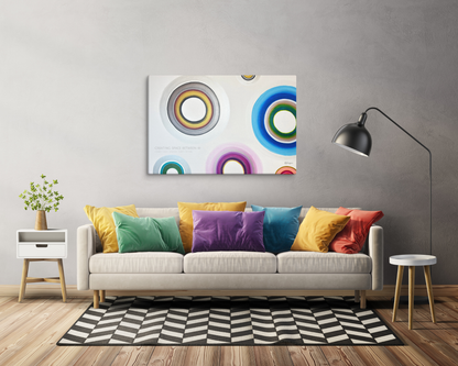 A modern living room scene featuring a canvas wall art print with colorful concentric circles, hung above a light beige sofa with vibrant multicolored throw pillows.